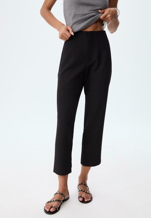Schwarze, hoch taillierte Hose mit geradem Schnitt und strukturiertem Stoff, ausgestattet mit vorderen Bügelfalten und einem glatten Bund. Kombiniert mit flachen Sandalen.