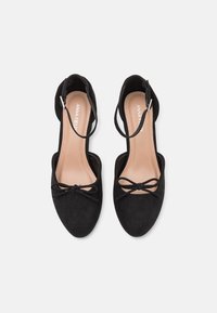 Paire de chaussures plates noires avec brides à la cheville et nœuds sur les orteils, semelles intérieures beiges imprimées du nom de la marque "Anna Field".