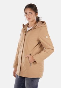 Giacca beige con cappuccio realizzata in tessuto liscio, dotata di zip frontale, due tasche laterali e una patche logo sulla manica sinistra.