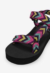 Sandalias coloridas con suela negra que presentan un diseño de tiras trenzadas en patrones de zigzag de color rosa, morado, azul verdoso y amarillo.