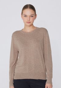 Jeune femme à la peau claire et aux cheveux attachés, portant un pull en maille beige et un pantalon noir, tournée vers l'avant sur un fond clair uni.