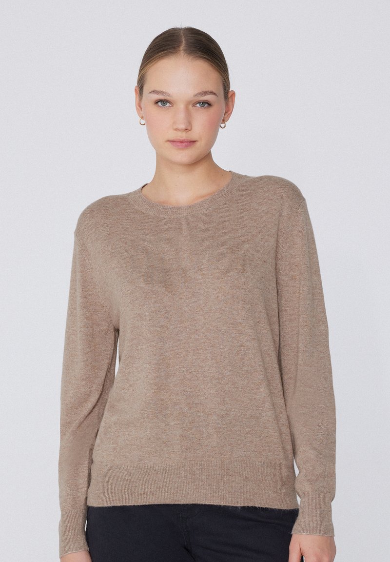 Jeune femme à la peau claire et aux cheveux attachés, portant un pull en maille beige et un pantalon noir, tournée vers l'avant sur un fond clair uni.