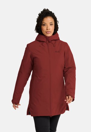Frau trägt eine lange weinrote Winterjacke mit Kapuze und Reißverschluss vorne sowie schwarze Hose, steht vor einem einfarbigen hellen Hintergrund.