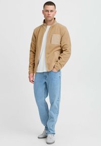 Tan fleece jacket with a zip, featuring a beige pocket. Worn over a white t-shirt, paired with light blue jeans and gray sneakers.