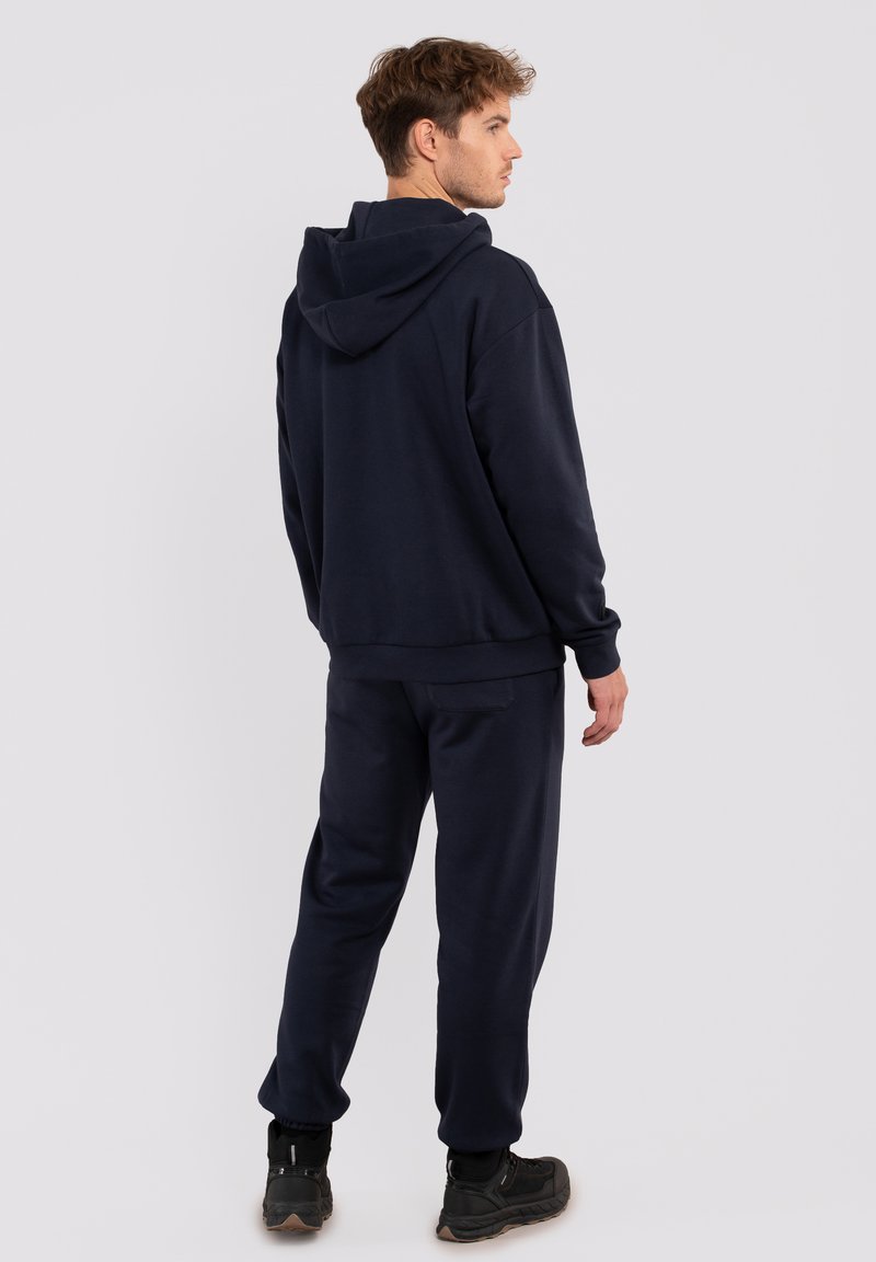 Mann in dunkelblauer Kapuzenjacke und passender Jogginghose steht, mit dem Rücken zu uns und schaut zur Seite vor einem schlichten hellen Hintergrund.