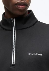 Κοντινό πλάνο ενός ατόμου που φοράει μαύρο τοπ Calvin Klein με φερμουάρ και λευκό λογότυπο στο στήθος.