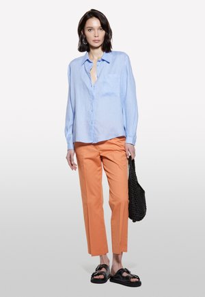 Chemise bleue claire à boutons avec une poche frontale, associée à un pantalon orange à jambes droites et des sandales noires. Un sac tissé noir complète le look.