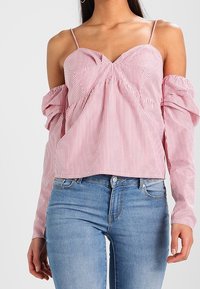 Punavalkoraidallinen olkapäät paljastava pusero, jossa ohuet olkaimet, poimutettu etusauma ja pitkät pörröiset hihat. Yhdistetty vaaleansinisten farkkujen kanssa.