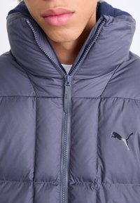 Giacca a vento grigia con colletto alto, chiusura con zip e logo riflettente. Presenta una texture trapuntata e un tessuto liscio.
