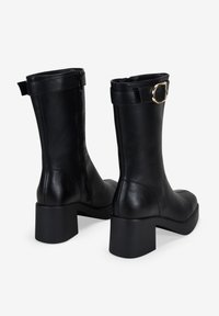 Botas de tobillo de cuero negro con un tacón grueso, cremallera lateral y un detalle de hebilla dorada en la correa superior. Textura suave. Diseño de punta cerrada.