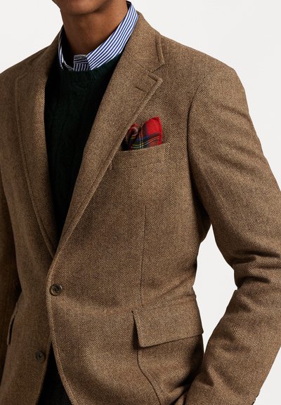 Bruine herringbone blazer met een notch lapel en twee knopen. Gecombineerd met een groene kabelgebreide trui en een blauw gestreept overhemd, met een rode geruite pochet.