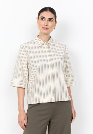 Woman wearing a cream blouse with thin vertical stripes and wide sleeves, paired with olive green pants, standing against a white background.