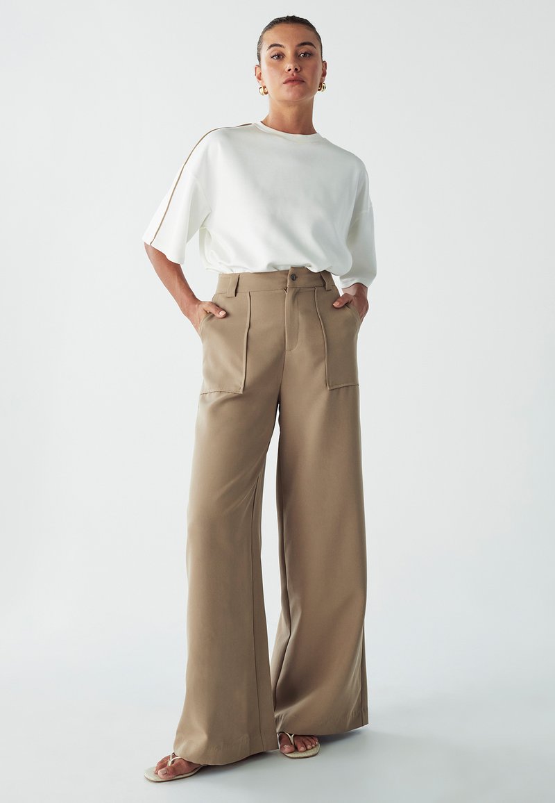 Pantaloni khaki a gamba larga con tasche frontali e vita aderente, abbinati a una maglietta bianca ampia con strisce color sabbia lungo le maniche.
