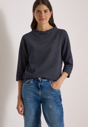 Jeune femme aux longs cheveux foncés portant un haut rayé bleu marine à manches trois quarts et un jean bleu, une main dans la poche.