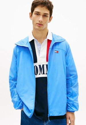 Joven con chaqueta azul claro con cremallera sobre una camisa polo de bloques de color blanco, rojo y azul marino con texto, posando frente a un fondo blanco.