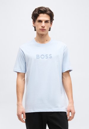 Giovane uomo con capelli castani mossi, indossa una maglietta azzurro chiaro con la scritta "BOSS" e pantaloni neri, in piedi davanti a uno sfondo chiaro e uniforme.