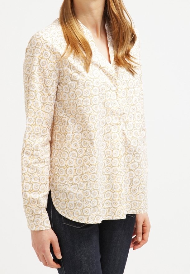 Blusa bianca a maniche lunghe con un motivo circolare dorato, dotata di colletto e fondo curvo, abbinata a jeans scuri.