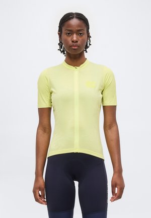 Femme aux cheveux tressés portant un maillot de cyclisme à manches courtes vert clair et un cuissard noir, debout sur un fond blanc.