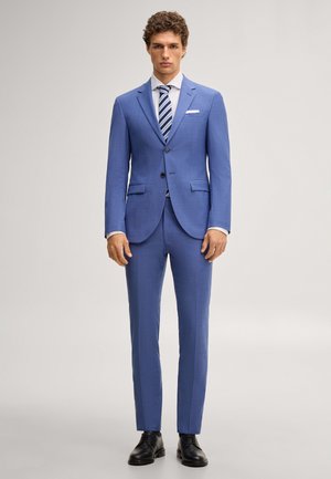 Mężczyzna w dopasowanym, jasnoniebieskim garniturze z białą koszulą, niebieskim paskowanym krawatem, białą poszetką i czarnymi eleganckimi butami, stojący twarzą do przodu.