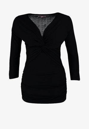 Mujer con top negro de manga larga y escote en V, jeans oscuros ajustados, botines negros con tachuelas, collar de cadena y bolso negro con correa de cadena.