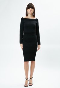 Schwarzes schulterfreies Kleid mit strukturierten Mustern, enganliegender Silhouette und knielangem Saum. Kombiniert mit schwarzen Riemchenschuhen.