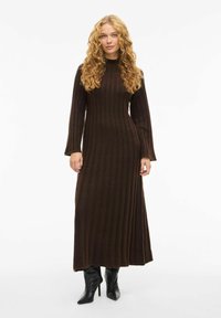 Robe en tricot côtelé marron avec des manches longues et un col haut, présentant un corsage ajusté et une jupe fluide. Portée avec des bottes à talons aiguilles noires.