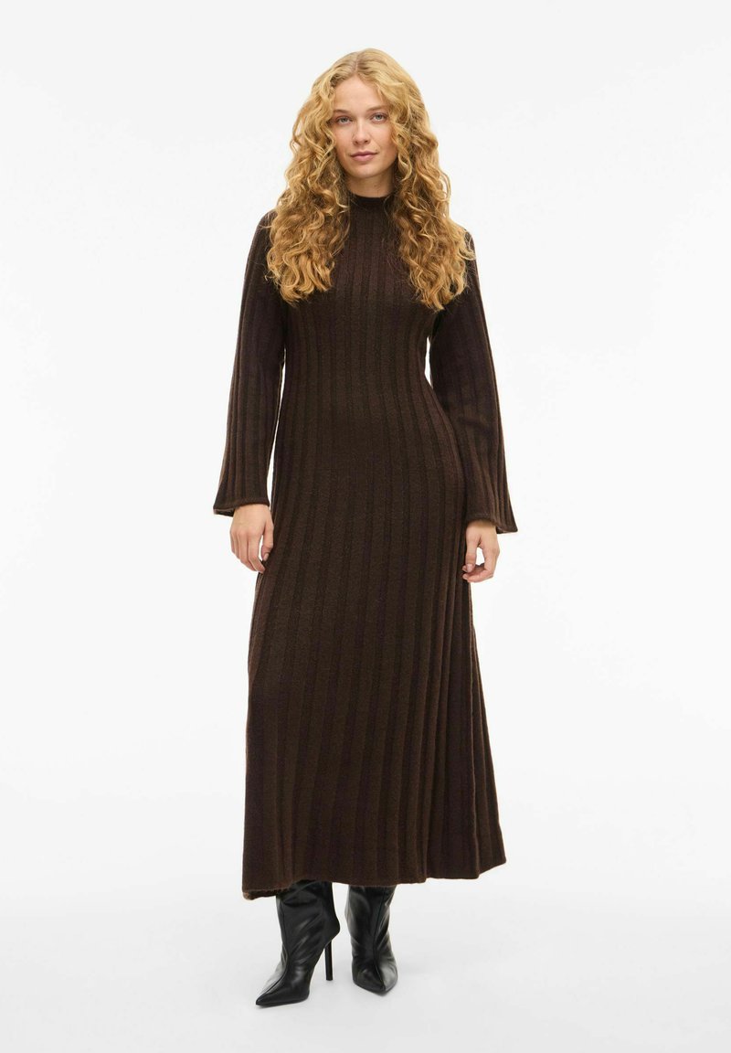 Robe en tricot côtelé marron avec des manches longues et un col haut, présentant un corsage ajusté et une jupe fluide. Portée avec des bottes à talons aiguilles noires.