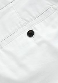 Gros plan sur un tissu blanc avec un bouton noir fermant une poche fendue horizontale, montrant les coutures détaillées et la texture.
