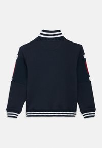 Μπλε ναυτικό φούτερ με ψηλό γιακά, ριγωτές λευκές και μαύρες ρίγες στα μανίκια και το τελείωμα, και έγχρωμες λεπτομέρειες στα μανίκια.