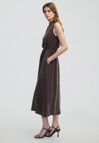 Hněd dress bez rukávů s vysokým výstřihem, stáhnutým pasem a kapsami. Hladká textura, s volným siluetou a délkou ke kotníkům.