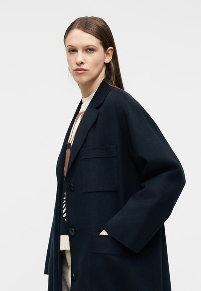 Γυναίκα με μακριά καστανά μαλλιά που φοράει ένα σκούρο ναυτικό oversized παλτό και ένα πουλόβερ με σχέδια, ποζάροντας μπροστά σε μια απλή γκρι φόντο.