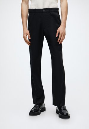 Persona che indossa pantaloni neri a gamba dritta, mocassini neri lucidi e un top chiaro senza maniche, in piedi con le braccia rilassate.