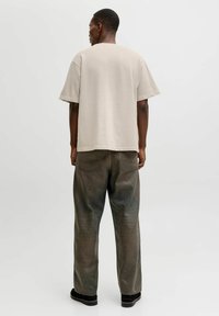 Moški obrnjeni stran, oblečen v bež preveliko majico, izprane črne široke kavbojke, črne čevlje in obročasto uhani v nevtralnem okolju.