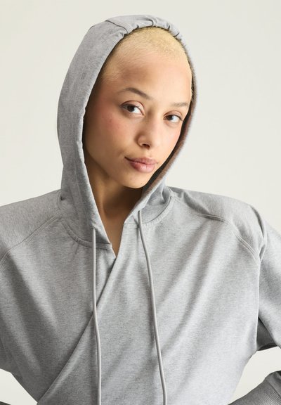 Person with short blonde hair wearing a gray hoodie with the hood up, looking directly ahead with a neutral expression.