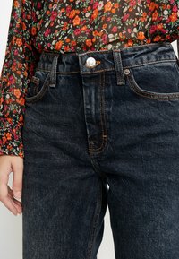 Jeans in denim scuro a vita alta con cuciture arancioni in evidenza, tasche anteriori e chiusura con bottone metallico. Tessuto testurizzato con leggero sbiadimento.