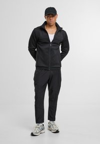 Černá zipová bunda s vysokým límcem, spárovaná s odpovídajícími zúženými kalhotami. Nošena s černou čepicí a stříbrnými sportovními botami s síťovanými detaily.