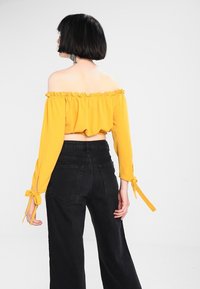 Gelbes, schulterfreies Crop-Top mit Rüschensaum und gebundenen Ärmeln, kombiniert mit hoch taillierten schwarzen weit geschnittenen Jeans.