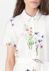 White short-sleeve dress shirt featuring a floral print in purple, pink, and red. Button-down front with a tied waist. Soft fabric texture.