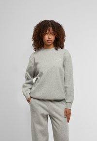 Pilkas džemperis su ilgomis rankovėmis, apvalia iškirpte ir raišteliais. Ant krūtinės yra mažas baltas logotipas, o medžiaga yra minkšta ir tekstūruota.