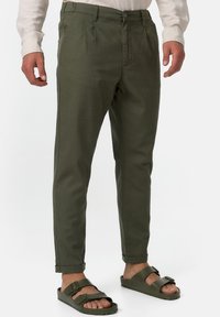 Khaki vihreät chinot, joissa on sileä pinta, kapeampi istuvuus, etupliitit ja rullatut hihansuut, yhdistetty tummanvihreisiin liukusandaaleihin.