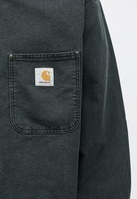 Τσέπη μαύρου σακακιού από κορντούρο με ραμμένη λευκή και κίτρινη ετικέτα λογότυπου Carhartt στο κέντρο.