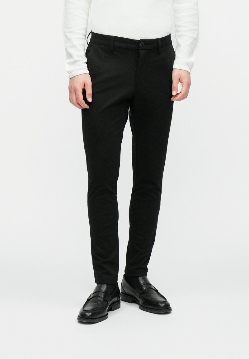 Muž v slim-fit čiernych nohaviciach, čiernych kožených mokasínach a bielej košeli s dlhým rukávom stojí proti jednoduchému pozadiu.