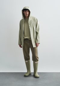 Oliwkowo-zielona kurtka przeciwdeszczowa, beżowy sweter, beżowe skrócone spodnie i zielone gumowe kalosze. Kurtka posiada kaptur i zapięcie na zatrzaski.