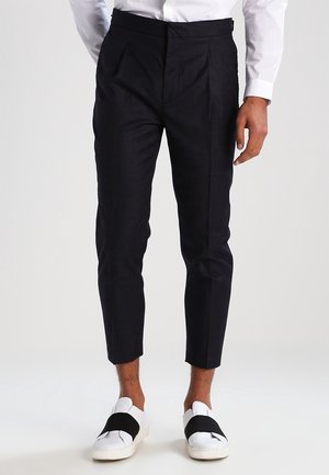 Mężczyzna w czarnych, skróconych, dopasowanych spodniach, białej koszuli i białych wsuwanych trampkach z szerokimi czarnymi paskami.