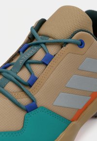 Pantofii sport bej au un superior texturat, cu accente turcoaz, sireturi albastre și dungi gri. Au vârful întărit și o talpă flexibilă.