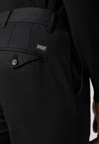 Zwarte broek met een gladde textuur, voorzien van een achterzak met een knoopsluiting en een label met de tekst "Scotch & Soda."