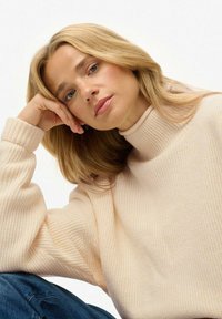 Femme blonde aux yeux bleus portant un pull à col roulé crème à côtes, reposant sa tête sur sa main, assise contre un fond blanc.
