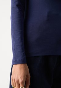 Marinblå långärmad skjorta med en slät textur, som har en figurnära design och fållade ärmslut. Dekorerad för hand med flera ringar.