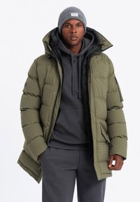 Chaqueta acolchada de color verde oliva con cierre de cremallera y botones a presión, usada sobre una sudadera gris; combinada con pantalones a juego y un gorro gris.