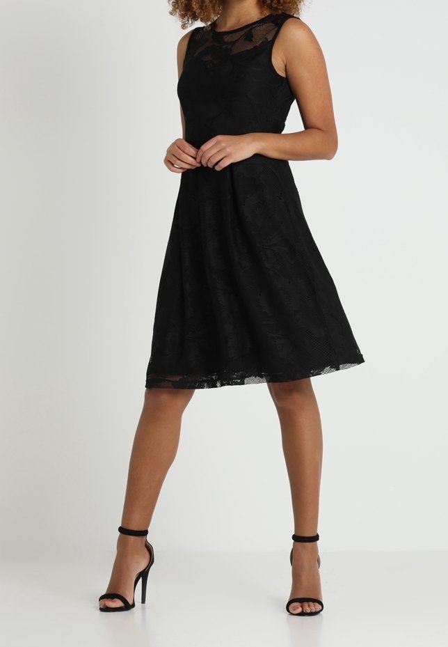 Femme debout portant une robe noire en dentelle sans manches jusqu'aux genoux et des sandales noires à talons hauts avec bride à la cheville sur fond blanc.